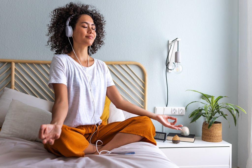 Young African American woman meditating at home sitting on the bed following online meditation, listening with headphones, using phone app. Technology and wellness concepts.