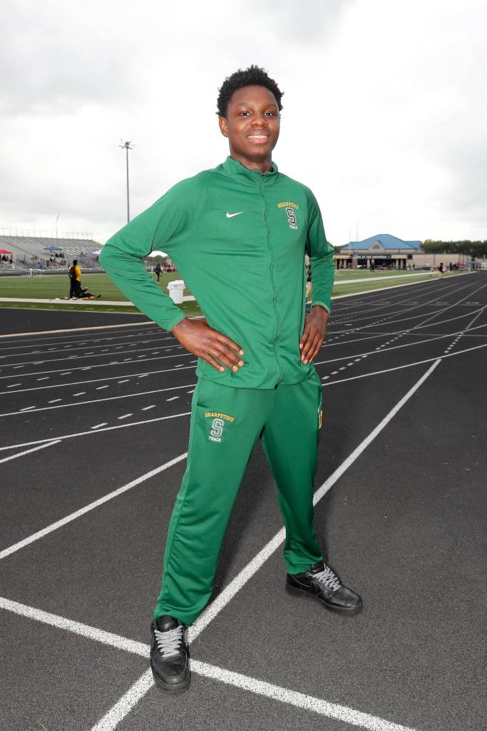 young Black teen poses outdoors on a track with hands grabbing each side of his waist, dressed in a green tracksuit